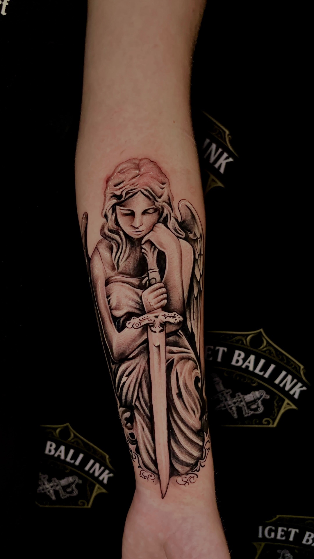 Angel statue with sword forearm tattoo in black and grey